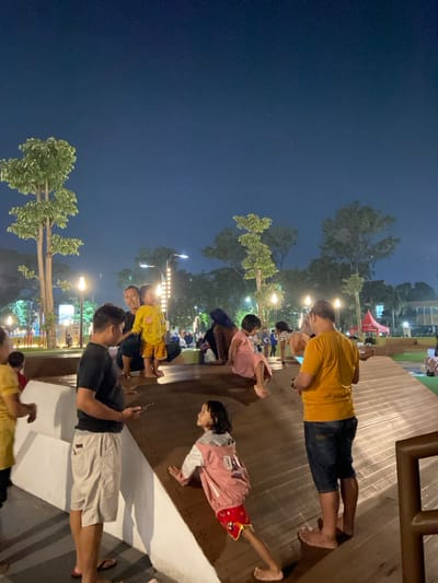 A nighttime scene is depicted in an outdoor public space in Sidoarjo, Indonesia, illuminated by artificial lighting from lampposts and decorative vertical light strips. The environment features multiple trees with green foliage and sloped wooden recreational structures. In the immediate foreground, a man in a black t-shirt and light-colored shorts, wearing flip-flops, stands on a flat white concrete surface, engaged with a smartphone. Adjacent to him, a young girl with dark hair, wearing a pink and white jacket over red patterned shorts, barefoot, is positioned on a sloped wooden surface, leaning forward. To the right, another man in a yellow t-shirt and dark blue shorts, barefoot, stands facing away from the camera on a sloped wooden ramp, holding a smartphone with both hands. In the mid-ground, a woman crouches on a flat surface, smiling towards the camera, alongside a small child in a yellow patterned outfit who is also looking towards the camera. Various other individuals, including adults and children, are visible throughout the scene, some sitting or playing on the wooden structures and grass areas. A red tent structure is discernible in the distant background on the right. The sky is dark blue, indicating night.