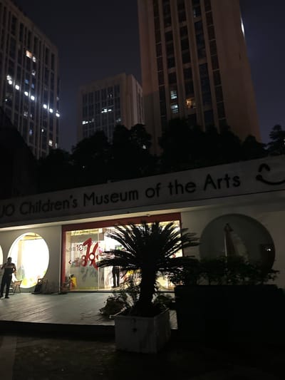 At night, an exterior view shows the entrance to the "Children's Museum of the Arts" in Chongqing High-tech Zone, China. The building is lit up with interior lights visible through large arched windows and glass doors. A sign above the entrance reads "Children's Museum of the Arts" with a smiley face logo. Inside the museum, displays of children's artwork and promotional materials are visible. A small palm tree stands in a planter to the right of the entrance. Two figures are seen near the entrance, one walking and one standing. In the background, tall buildings are illuminated against the dark sky.