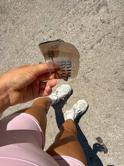 A pair of hands holds a pair of sunglasses with "MIU MIU" embossed on the lenses. The sunglasses are transparent with a metallic logo. The background shows a person's legs wearing light pink athletic shorts and white sneakers with white socks. The scene is set on a grey, textured surface, likely concrete or pavement. The lighting suggests it is daytime, with shadows indicating a sunny atmosphere. The overall mood appears casual and perhaps active, given the athletic wear. The location is identified as Fethiye, Turkey.