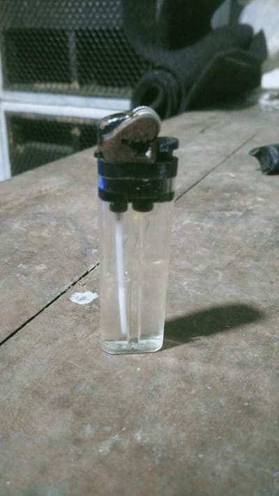A disposable cigarette lighter, filled with clear liquid fuel, sits on a rough, weathered wooden surface. The lighter is translucent, revealing the liquid inside, and has a black plastic top with a metal sparking mechanism. The background is out of focus, showing what appears to be a metal grate structure and a dark, rolled-up object. A shadow is cast by the lighter onto the wooden surface. The location is the city of Wonosegoro, Indonesia.
