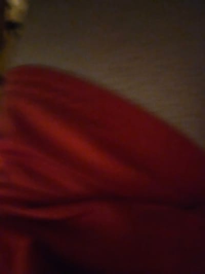 The image displays a close-up, out-of-focus view of a draped red fabric. The lighting is dim and appears to be coming from the side, creating subtle highlights and shadows that suggest folds and texture in the material. The background is dark and indistinct, with a slight gradient of muted brown and grey tones. There are no discernible people, objects, or environmental features that would indicate a specific location within the city of Versailles, France. The image is primarily a study of texture and color in a deep red hue.