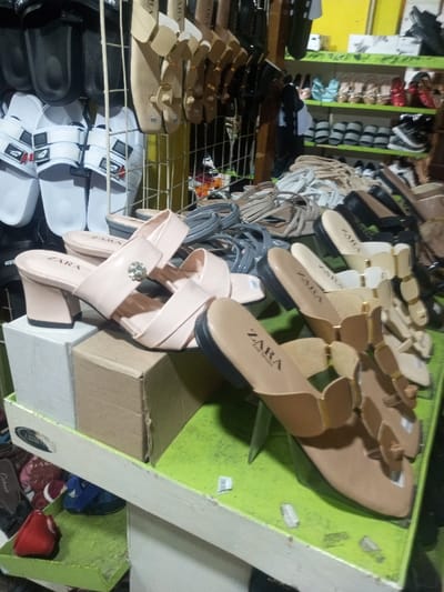 A variety of sandals and shoes are displayed on shelves and a table. Black and white slides hang above. Below, a selection of strappy sandals and ZARA branded slip-on shoes are neatly arranged. A pair of pink heeled sandals rests on stacked boxes in the foreground. The items are well-lit, suggesting an indoor retail setting.