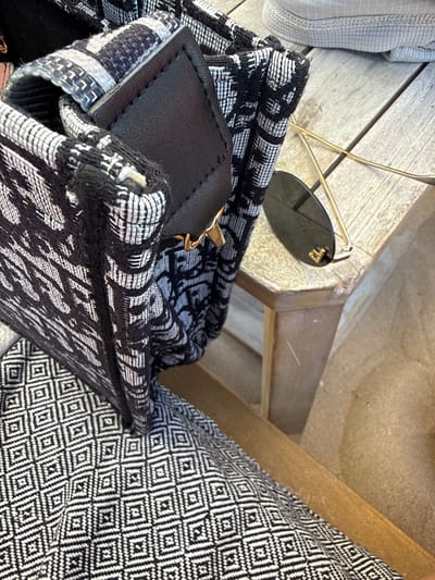 The image shows a close-up of a patterned textile bag, possibly a tote bag, with a black leather detail and a gold clasp. A pair of gold-rimmed sunglasses with dark lenses rests on a weathered wooden surface, likely a table or bench. The background is blurred, but suggests an outdoor setting. There are no people visible in the image. The scene is set in Nesebar, Bulgaria.