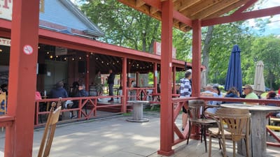 This image captures an outdoor dining area with a red wooden structure that appears to be a covered patio or veranda. The structure has a wooden roof supported by red posts and beams. String lights are suspended from the ceiling, indicating it is likely an evening or a dimly lit setting, though the bright daylight suggests otherwise. Several tables and chairs are arranged on the patio and in the adjacent area, with some occupied by people.
In the background, a brick building with a light-colored roof and red trim is visible. A stage setup with microphones and speakers suggests a live music performance may be taking place or has recently concluded. Signs are posted on the building, one of which appears to be a "no smoking" symbol and another with text that includes "Desjardins."
The environment is leafy and green, with trees providing shade and a backdrop of foliage. The ground beneath the patio is paved with light-colored tiles or concrete. The overall scene suggests a casual, social gathering place, possibly a restaurant or bar. The presence of people engaged in conversation and enjoying the outdoor setting points to a relaxed atmosphere.
While the specific location in Pointe-des-Cascades, Canada, cannot be definitively determined from visual cues alone, the style of the building and the general ambiance are consistent with a Canadian setting, particularly during warmer months. The presence of a "Desjardins" sign indicates a connection to a Canadian financial institution.