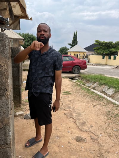A full-body, medium shot captures a Black man with a beard standing outdoors, facing forward and slightly to the left, in what appears to be a residential area in Abuja, Nigeria. He is wearing a short-sleeved, black collared shirt with a gray leaf pattern and black shorts. His left hand is raised to his chin, and his right hand is by his side, holding a black object. He is wearing black slide sandals. The man stands on a dirt path next to a rough, concrete structure. In the background, a red sedan is parked on a paved road. Beyond the road, there are yellow and light-colored buildings, some with pitched roofs, and lush green trees. The sky is overcast with gray clouds. A narrow, vegetated drainage ditch runs along the side of the road. The overall impression is a casual, everyday scene in an urban environment with modest infrastructure.