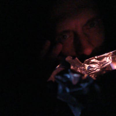 A person with fair skin holds a crinkled piece of foil near their mouth, appearing to be smoking from it. The scene is dimly lit, possibly at night. The person’s face, particularly the eyes and nose, is visible. The location is not discernible from the visual information provided.