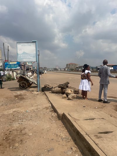 The image displays an urban street scene in Kwashieman, Accra, Ghana, identifiable by a prominent sign reading "CHRIST APOSTOLIC CHURCH INTERNATIONAL KWASHIEMAN ASSEMBLY." The foreground features a rough, unpaved dirt shoulder with scattered debris, transitioning into a damaged concrete curb and an exposed drainage channel.

To the left, a blue-framed sign stands erect, positioned next to a discarded, rusted metal pushcart or wheelbarrow that appears partially loaded. Immediately to the right of the sign, a pile of large rocks, several cut logs (possibly palm tree trunks), and a single black tire are stacked on the dirt ground.

On the right side of the frame, two individuals stand on the dirt shoulder. A woman, dressed in a white sleeveless dress and flat sandals, carries a small child secured on her back with a patterned fabric wrap. She is observed looking towards the paved road. Adjacent to her, a bald man wearing a patterned grey and white suit and dark shoes stands with his back mostly to the camera, extending his right arm and hand in a gesturing motion towards the street.

In the mid-ground, a multi-lane paved road extends into the distance, with several vehicles and pedestrians visible. Streetlights line the road, and a speed limit sign indicating "50" is discernible. The background features various commercial or residential buildings, including a multi-story structure on the far side of the road. The sky above is heavily overcast with dense grey clouds. A person in a green outfit is partially visible on the far left, near the pushcart.