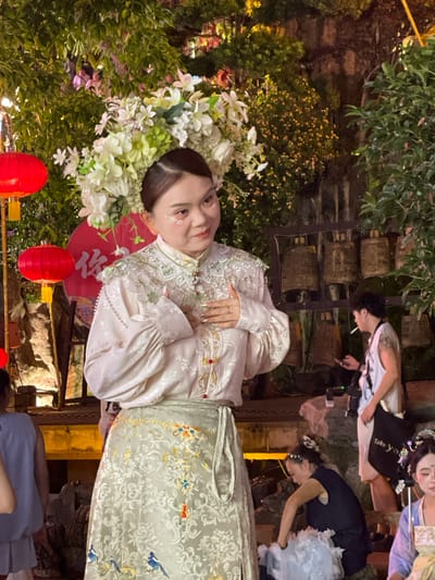 The image depicts a woman in a traditional Chinese dress with elaborate floral headwear, likely taken during a cultural event. Her attire is pale, adorned with embroidery and embellishments. She holds a decorative fan and is positioned among red lanterns and greenery. Other people are also in the vicinity, possibly staff and visitors.
The location is Yuzhong District, China. The scene appears to be outdoors.