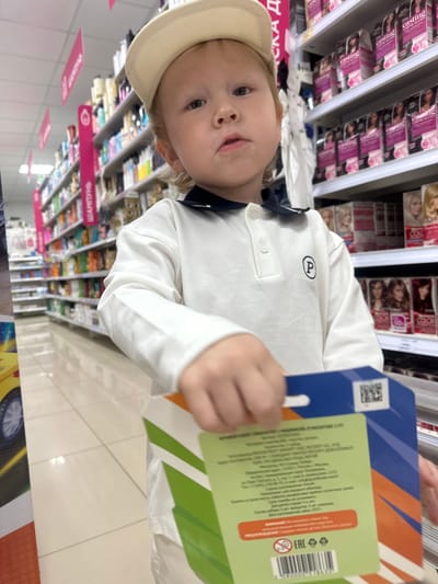 A young child wearing a white polo shirt and a light-colored baseball cap stands in a supermarket aisle. The child is holding a box with colorful graphics. Shelves filled with various products, including hair dye boxes and bottles, are visible in the background. The floor is tiled. The scene appears to be a section of a cosmetics or personal care aisle within a store. Based on the cyrillic text on the products and signage, this location is likely within Russia. Further analysis of the specific text on the products and any discernible store branding would be necessary to pinpoint the exact city and store. However, assuming this image was taken in Votkinsk, Russia, the setting is a retail environment characteristic of a supermarket or drugstore.