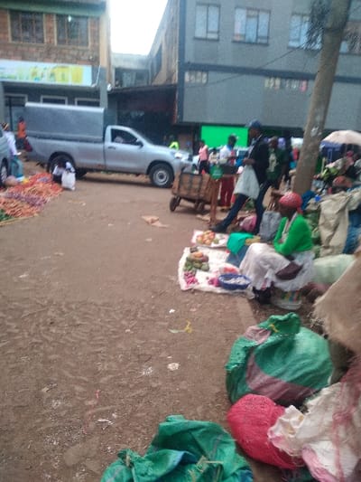 A street scene features a light-colored pickup truck parked near a building. People are visible in the background near a large green screen. A variety of sacks and bags in green, white, and red hues are scattered on the ground. A patterned cloth displays what appears to be fruits or vegetables. The ground is earthy in color and shows signs of wear.
