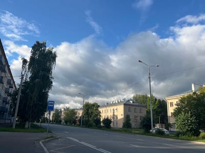 The image depicts a street scene in Kazan, Russia, under a partly cloudy sky. A road with clear lane markings runs through the center of the frame, bordered by grassy verges and sidewalks. To the left, a multi-story apartment building with visible balconies is partially visible. A tall, slender tree with drooping foliage stands near the sidewalk, adjacent to a blue bus stop sign. Several other apartment buildings, characterized by their light yellow brick facades and numerous windows, line the opposite side of the road. Green trees and bushes are interspersed among the buildings. Streetlights are present along the roadside. A white car is parked on the far side of the road. The lighting suggests it is daytime, with sunlight partially illuminating the scene, creating shadows and highlighting the texture of the buildings and clouds.