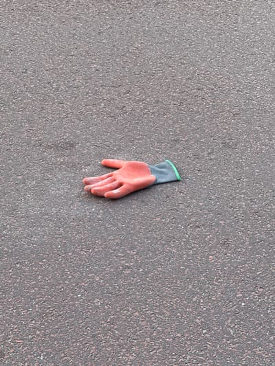 The image shows a single, left-handed gardening glove lying on an asphalt surface. The glove is made of a textured, coral-pink material, likely rubber or a similar synthetic, with a gray fabric cuff that has a thin, light green trim. The fingers of the glove are slightly curled, giving the impression of a relaxed hand.
The surface it rests on is a textured, dark gray asphalt, typical of a road or walkway. The texture of the asphalt is granular, with small pebbles and bits of aggregate visible. The lighting suggests it is daytime, with a soft, even light that casts no strong shadows, implying an overcast sky or perhaps a shaded area.
The scene is devoid of any other objects or people, placing sole focus on the discarded glove. The lack of context leaves the reason for its presence and abandonment to speculation. There is no visible text within the image. The overall impression is one of a mundane, perhaps slightly melancholic, scene of urban detritus.