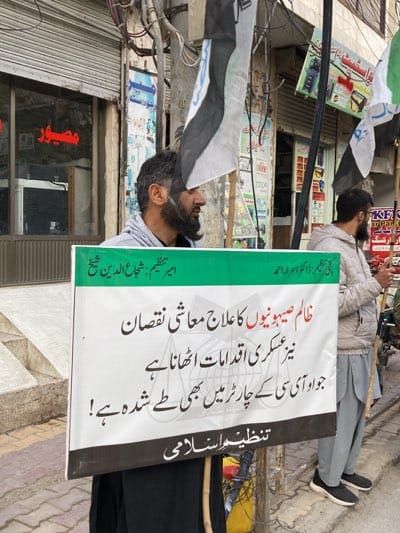 A medium, eye-level shot shows two men protesting on a street in Quetta, Pakistan.
The man on the left carries a white protest sign that is about three-quarters visible; it features black writing, mostly in Urdu. At the top it reads, "امیر تنظیم : شجاع الدین شیخ" ("Amir Tanzim: Shuja-ud-Din Sheikh") and "ظالم صیہونیوں کا علاج معاشی نقصان" ("The treatment of the cruel Zionists is economic loss"). The man holds a white and black flag aloft; he is bearded and light-skinned. The man on the right is similarly bearded and fair-skinned. He is wearing a grey windbreaker and is carrying a wooden staff. The two men stand on a street with sidewalks, power poles, and stores behind them. A sign above one of the stores reads "مصيور" (likely “Masir”). Flags are visible to the right of the man on the right.