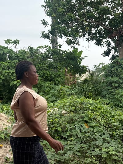 This image captures a woman standing outdoors in a lush, green environment in Ikot Ekpene, Nigeria, during the daytime under bright, diffused light.

The primary subject is a woman with dark skin and braided or dreaded hair pulled back. She is wearing a light-colored (beige or light brown) sleeveless top and dark pants with a light-colored checkered pattern. She is seen in profile, looking upwards and slightly to her left, with a neutral and calm expression. Her left arm is slightly bent and extended, with her hand open as if observing something or gently reaching.

The scene is dominated by dense, tropical vegetation. On the left, a large tree with abundant dark green leaves extends towards the top of the frame. The right side and background feature a variety of green plants, including broad-leafed plants that resemble cassava, intertwined vines with smaller leaves, and hints of palm fronds in the distance. The ground immediately around the woman is light-colored, appearing sandy or dusty, with some scattered yellow plant debris. The sky, visible through gaps in the foliage, is a bright, uniform white, suggesting an overcast day. No visible text is present in the image.