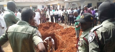 This image captures an outdoor scene in Umu Enge, Nigeria, centered around a large, freshly dug pit of reddish-brown soil. The primary activity appears to be the digging of this pit, possibly a grave, as a man in a light-colored shirt is visible inside it, actively using shovels.
A considerable crowd of people is gathered around the pit, observing the activity. Among them are several individuals in light olive-green uniforms with shoulder boards and patches. One prominent patch clearly displays "S.Q.B. NIGERIA" along with an emblem. Another uniformed person wears a black baseball cap featuring the yellow letters "SF." A person in uniform to the right is seen wearing a black balaclava. The rest of the attendees are dressed in a mix of attire, including white robes, suits, shirts, trousers, and patterned dresses, indicating a formal or communal gathering.
The setting is outdoors under bright, natural daylight, suggesting clear weather conditions. In the background, parts of white or light-colored buildings with tiled roofs are visible. The reddish-brown soil is a notable characteristic of the ground. The overall atmosphere conveyed by the gathering around the pit suggests a solemn event, likely a funeral or a burial ceremony.