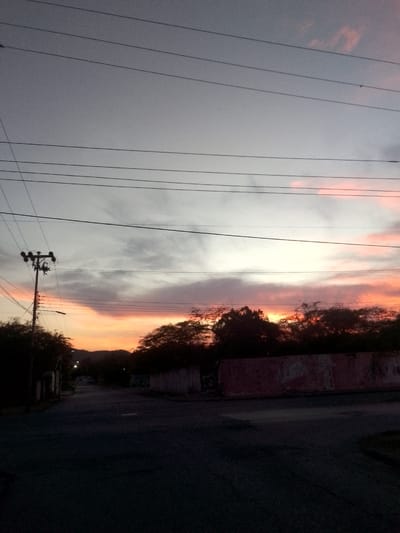 The image depicts a sunset over a street in Juan Griego, Venezuela, viewed from a low angle. The sky is a prominent feature, showing dark grey and light grey clouds at the top, transitioning to vibrant orange and pink hues along the horizon, indicating dusk. Several parallel power lines stretch across the upper and middle portions of the sky. To the left, a tall utility pole with multiple crossarms and power lines is silhouetted against the colorful sky, casting a long shadow.
The foreground and mid-ground elements are heavily shadowed due to the low light, appearing mostly in silhouette. Dark trees and dense foliage line the mid-ground, obscuring the horizon in places. To the right, a long, low wall with faded red and white paint is visible beneath the trees. A dark, unlit asphalt road occupies the foreground, extending into the distance where faint outlines of distant hills or mountains are barely visible against the twilight sky. Faint suggestions of streetlights or buildings are discernible along the road in the distance.
No people are present, and no specific activity is taking place, contributing to a serene atmosphere. The notable details include the striking sunset colors, the intricate network of utility lines, and the stark contrast between the bright sky and the darkened landscape. There is no visible text in the image. The scene evokes a sense of tranquil twilight in a residential or semi-rural setting.
