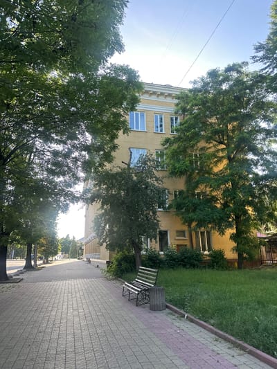 The image depicts a paved sidewalk in the city of Ternopil, Ukraine, bordered by a grassy area on the right and trees on the left. A yellow building with multiple windows stands behind the grassy area. A metal bench and a concrete trash can are positioned on the sidewalk. Sunlight casts shadows across the scene. The sky is clear and blue.