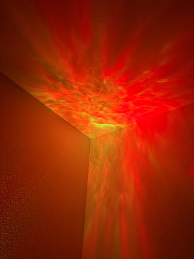 The image displays an interior corner of a room located in Gainesville, United States of America. A textured ceiling and two adjacent textured walls are visible. All surfaces are illuminated by projected light, featuring prominent red coloration combined with swirling, wave-like patterns of green light. These light patterns cover the entirety of the visible ceiling and extend down both visible walls. No individuals, discernible objects, or specific actions or interactions are present in the scene. The environment is an enclosed indoor space, defined by the extensive colored light projection.