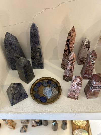 The image shows a collection of polished gemstones displayed on a white shelf. The gemstones are primarily in the shape of obelisks and pyramids, with some smaller tumbled stones. There are two main types of gemstones visible: a dark blue stone with white veining, and a reddish-brown stone with white and black inclusions, resembling jasper or agate. The blue stones are arranged on the left side of the shelf, with a pyramidal stone in the foreground and several obelisks behind it. The reddish-brown stones are on the right side, also displayed as obelisks of varying sizes. In the center of the shelf, there is a small, ornate brass dish containing several small, dark blue, oval-shaped stones and a larger, rougher piece of a similar blue stone. The background is a white wall with a visible crack running across the top. The shelf itself is also white and appears to be made of wood. There are no people visible in the image. This arrangement of polished gemstones is typical of those found in tourist shops or gemstone galleries, commonly sold in areas like Sukawati, Indonesia, which is known for its art markets and artisanal crafts.