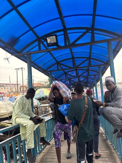 This image depicts a segment of a covered pedestrian walkway or bridge in Lekki, Nigeria, specifically above or adjacent to a bustling market area. The walkway features a blue translucent corrugated roof supported by a dark metal arched framework and blue metal railings. An overhead floodlight is mounted on the roof structure.

In the left foreground, a male individual wearing a light green traditional outfit (tunic and trousers) is seated on the blue railing, facing right, looking down at a smartphone held in his hands. His feet are bare.

Mid-ground, moving from right to left, a male individual with dark skin wears a black t-shirt and purple patterned shorts. He carries a large white and red striped sack, marked "KGI", over his right shoulder and a blue plastic bag in his left hand. He wears dark clogs. Directly behind him, another male individual is seen from the back, wearing a dark green t-shirt, black Puma track pants, and a brown crossbody bag. He also holds a blue plastic bag.

On the right mid-ground, a male individual wearing a light grey traditional outfit and a white patterned kufi is perched on the blue railing, facing forward. His right hand is near his chin, and a gold watch is visible on his left wrist. He wears dark sandals.

Additional male figures are partially visible further down the walkway, indicating ongoing foot traffic.

The background, visible beneath and beyond the walkway, reveals a vibrant market scene with numerous stalls, white and light-colored tarpaulin coverings, and various merchandise. Power lines, utility poles, and multi-story buildings, including one partially visible sign for "ALEST HOTEL", indicate an urban environment. The lighting suggests daytime conditions.
