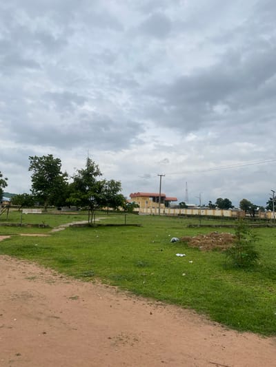 The image shows an outdoor scene in Jalingo, Nigeria, with a wide expanse of green grass in the foreground and middle ground, bordered by a dirt path on the left. A pile of dried vegetation and some scattered litter are visible on the grass. In the background, a modern, light-colored building with a red roof stands behind a beige and white fence. Several trees, some tall and leafy and others smaller, are scattered across the landscape. A telecommunications tower is visible in the distance to the right of the building, and power lines stretch across the sky. The sky is overcast with heavy, gray clouds, suggesting a potentially dull or rainy day. No people or animals are present in the image. The overall impression is one of an undeveloped or semi-developed urban area.