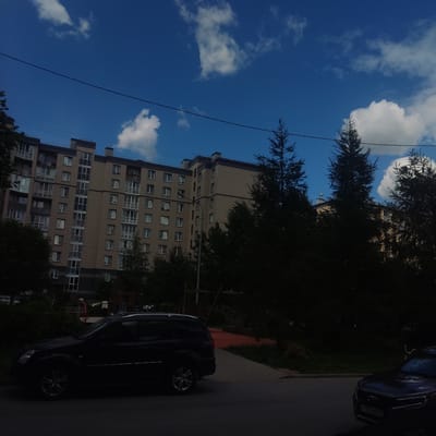 A wide-angle, low-angle shot captures a blue sky with scattered white clouds above a multi-story residential building. The building appears to be made of beige and gray materials, with numerous windows and balconies. Trees with dark green foliage are interspersed between the building and the sky, and in the foreground. A dark-colored car is parked on the right side of the image, facing away from the viewer. A sidewalk runs alongside the car. The image has a slightly tilted perspective, as if taken from ground level looking upwards. The lighting suggests it is daytime, possibly late afternoon given the shadows. A single power line runs diagonally across the frame. A license plate on the car is partially visible and appears to read "Y 137 AM 198". The scene depicts an urban or suburban residential area.