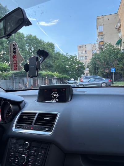 The image shows the interior of a car, viewed from the driver's perspective, with a dashboard display showing the Opel logo, the time 19:47, and the date 11.06.2026, with a temperature of 23°C. A car air freshener shaped like a shoe with the brand "SHOPSECTOR" is hanging from the rearview mirror. A phone holder is attached to the windshield. Outside the car, trees, apartment buildings, and parked cars are visible. A street sign with a blue background and a white arrow pointing down is seen on the right side. The scene suggests a sunny day in a residential area, possibly in the city of Panagyurishte, Bulgaria.