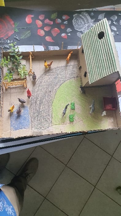 The image displays a handcrafted diorama of a chicken farm, situated on a dark surface decorated with painted red rose petals, green leaves, and white rose outlines. The diorama is constructed primarily from cardboard. Within the enclosure, a multi-story chicken house made of striped green cardboard with three visible circular openings is positioned in the upper right. The floor of the diorama is segmented: a light brown-grey area, scribbled to represent dirt or a path; a green colored area simulating grass; and a blue colored area depicting water. A total of nine small plastic chicken figures, exhibiting various colors including red, yellow, black, brown, and orange, are dispersed across these ground sections. Two green rectangular feeders and one red rectangular feeder are present, with a small white irregular object near the red feeder. Additional structures include a small yellow wooden fence or bench in the upper left, and a wooden swing-like perch. An artificial green leafy plant cluster is located near the fence. The bottom left of the image displays a light grey tiled floor with dark grout lines, partially occupied by the lower left leg and foot of an individual wearing a light blue patterned garment and a light brown shoe. This scene is located in Kisumu, Kenya.