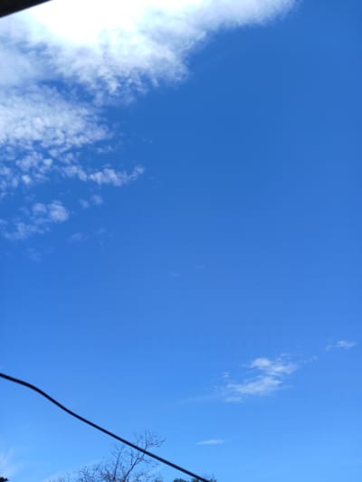 The image shows a bright blue sky with scattered white clouds. The sky is the dominant subject, taking up most of the frame. On the left side, there is a large mass of bright white clouds, possibly illuminated by the sun. Further into the sky, there are smaller, wispy clouds. The bottom right corner of the image reveals the bare branches of a tree and a dark, curved line which appears to be a wire.
The scene suggests an outdoor setting on a clear day. The presence of clouds indicates it is daytime, and the brightness implies fair weather. The limited visible elements and the angle of the shot suggest the photo was taken looking upwards. The location context, Altagracia, Venezuela, places this scene in a tropical or subtropical region, which aligns with the bright sky and cloud formations. There is no discernible activity or event taking place; the image is a snapshot of the sky. No text is visible in the image.