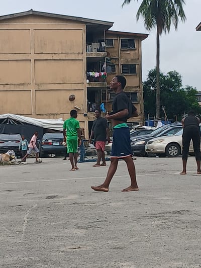 The image captures a street scene in Lagos, Nigeria, focusing on a group of young men playing football. The setting appears to be an urban residential area, with a multi-story, weathered building dominating the background. The building's facade is a light brown color, with visible signs of age and wear, and laundry is hanging from some of the balconies. Adjacent to the building, there's a collection of parked cars, including a dark sedan and a silver sedan. The ground is a paved asphalt surface, typical of streets or open areas in Nigerian cities, with some visible cracks and patches.

In the foreground, a young man in a black t-shirt and blue shorts is the most prominent figure, captured mid-motion, possibly looking upwards or in anticipation of the ball. To his left, a young man in a bright green t-shirt and matching shorts is also in motion, appearing to be involved in the game. Further to the left, a younger boy in a light blue t-shirt and shorts is kicking a football, indicating the game is actively underway. Another man in a black t-shirt and pink shorts is standing nearby, observing the game. To the far right, a person in a black t-shirt and dark pants is facing away from the camera, bent over, possibly participating in or watching the activity. The overall atmosphere suggests a casual, informal sporting event taking place in a communal urban space. A tall palm tree is visible to the right of the building, adding a distinctly tropical element to the Lagos scenery.