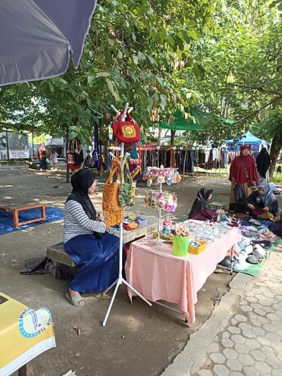 This vibrant image captures an outdoor market scene in Mataram, Indonesia, under the bright daylight, likely in a park or community area shaded by large trees. The ground is a mix of concrete and paving stones.
Numerous people, predominantly women wearing hijabs, are engaged in market activities. In the foreground on the left, a woman in a striped shirt, blue skirt, and black hijab sits on a concrete bench, seemingly tending her stall. Beside her, a white, multi-tiered stand displays an assortment of crocheted or handcrafted bags, including a prominent red one with a yellow flower pattern and a textured orange bag with fringe. Next to this, a table draped in a light pink cloth showcases various small accessories, such as keychains, hair clips, and floral decorations, arranged in baskets and on display stands. Another woman in a dark hijab is visible behind this table, focusing on the merchandise.
Further to the right, two other women are involved in sales. One, dressed in a striking red ensemble, stands, while another, also in a dark hijab, sits on the ground, arranging clothes and shoes laid out on a green mat. In the mid-ground, numerous clothing racks are visible, covered by green tarpaulins, indicating more stalls selling garments. A large gray umbrella on the far left provides additional shade. In the background, beyond the stalls, other people, including children, can be seen walking around, suggesting a lively community gathering.
Notable text includes a sign in the background reading "WARUNG MIE," followed by "MENU," "MIE AYAM," and "MIE GORENG," indicating a food stall. On a yellow surface in the bottom left, partial text "BALOM" and "-FEB" is faintly visible. The scene conveys a busy, localized marketplace atmosphere.