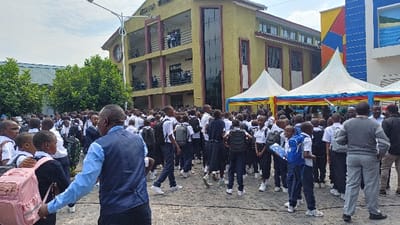 The image captures a bustling outdoor scene at the INSTITUT PALAIS DE GOMA in Goma, Democratic Republic of the Congo. Approximately 150-200 individuals, primarily students, are gathered on paved grounds under a partly cloudy sky.

The prominent structure in the background is a three-story building, painted yellow-ochre, featuring dark balconies with railings and large vertical dark glass panels. The inscription "INSTITUT PALAIS" is visible on its upper facade. To the right of this main building, a second multi-colored structure is visible, predominantly blue with abstract geometric patterns in yellow, red, and black, and a mural depicting a seascape.

In the midground, several white canopy tents with colorful red, yellow, and blue fabric draping are erected, indicating an event or gathering.

The majority of the people are young students, uniformed in white short-sleeved shirts and dark trousers or skirts. Many carry backpacks of various colors, including pink, blue, grey, and black. They are observed standing, walking, and interacting in groups across the central and left areas of the paved courtyard. Several adults are interspersed among the students and at the edges of the crowd. One adult male is in the foreground, facing away from the camera, wearing a light blue shirt and a dark vest. Another adult male stands on the right, wearing a grey sweater with striped sleeves and light grey trousers. A street light fixture on a pole is visible on the left side of the frame. The ground is consistently paved with interlocking stone pavers.