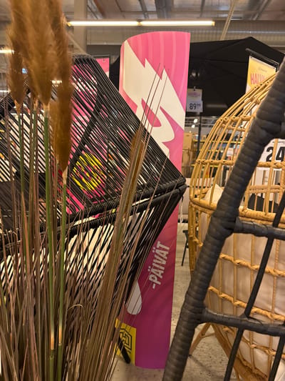 The image shows an indoor retail setting, likely a department store or furniture store, with a focus on decorative items and furniture. In the foreground, a large arrangement of dry, tall grass or reeds is prominent, with some stalks showing brown and green hues. Behind the grass, a modern black, geometric-shaped chair with a woven design is partially visible. To the right of this chair, a wicker hanging chair with a light-colored cushion is displayed.
A tall, pink and white vertical banner is positioned behind the black chair, featuring abstract shapes and text in white. Part of the text visible is "-PÄIVÄT". Further in the background, a yellow banner with the text "HALVA" is visible above a black umbrella structure. Numbers "24" are also visible on a white price tag. The overall environment suggests a well-lit retail space with a focus on home furnishings. The mention of Pori, Finland, would suggest this is a specific store within that city.