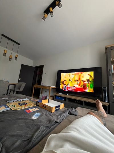 The image depicts a living room scene. A large television screen displays a news program with Bulgarian text. On the floor in the foreground, a board game, likely Monopoly, is set up with game pieces and play money scattered on a grey blanket. A person's bare leg, with tattoos, is visible on the right side of the frame, indicating someone is relaxing on a couch. In the background, there is a dining table with chairs, a doorway leading to another room, and a bookshelf. The lighting suggests it is daytime. The overall impression is of a casual home environment. There is no specific indication of the location being Vratsa, Bulgaria.