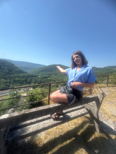 A woman stands on a stone platform overlooking a lush green valley under a clear blue sky. She is wearing a blue short-sleeved shirt and a black leather skirt. Her right arm is extended, and she is looking towards the camera with a smile. The scene appears to be set on a sunny day, likely in the late morning or early afternoon given the bright sunlight and long shadows. The location is identified as Lavans-lès-Saint-Claude, France, which is suggested by the hilly, forested landscape. The platform has a metal railing and is made of rough-hewn stone. The grass and dry vegetation on the platform suggest a natural, outdoor environment.