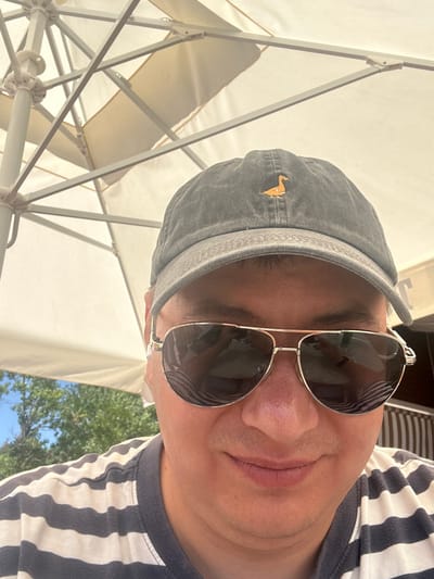 A close-up, self-portrait style image shows a person wearing a gray baseball cap with a small, orange duck embroidered on the front. They are also wearing aviator-style sunglasses with silver frames and dark lenses, and a navy and white striped t-shirt. The person is outdoors, with a large, cream-colored umbrella providing shade in the background. Trees and a bright blue sky are visible beyond the umbrella. The overall setting suggests a sunny day, likely in an outdoor dining or seating area, potentially at a restaurant or cafe. The location is specified as Prijevor, Montenegro.