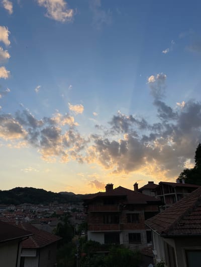 The image depicts a sunset over the city of Zlatograd, Bulgaria. The sky is a mixture of blue and golden hues, with scattered clouds catching the last rays of sunlight. In the foreground, several houses with tiled roofs are visible, some appearing to be under construction. A dense green hillside with more houses and trees forms the backdrop to the city, with mountains further in the distance. The overall scene conveys a tranquil and picturesque view of a European town at dusk.