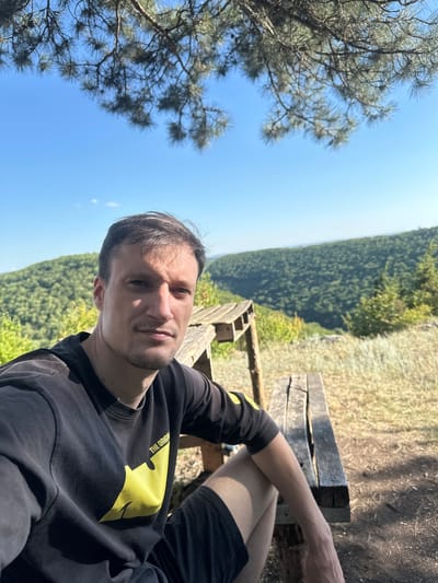 A young man with short brown hair and a stubble is taking a selfie outdoors. He is wearing a black sweatshirt with a large yellow graphic on the front and black shorts. He is sitting on a wooden bench outdoors, with a view of a forest and a valley behind him. The sun is shining, and the sky is clear blue. The image appears to have been taken during the daytime. The man's expression is neutral, and he seems relaxed.
The man is the primary subject. The wooden bench he is sitting on, a pine tree in the foreground, and the forest and valley in the background are other objects and scene elements. The activity taking place is a selfie being taken by the man.
Notable details include the clear blue sky, suggesting fair weather. The lush green forest and the sloping terrain of the valley hint at a natural, possibly mountainous or hilly, outdoor setting. The time of day is likely daytime due to the bright lighting and shadows. The man's relaxed posture and neutral expression suggest he is enjoying his time outdoors. The yellow graphic on his sweatshirt appears to contain text, possibly "THE ORIGIN," though it is partially obscured.
The image context provided is Velykozalissia, Ukraine. This suggests the photo was taken in that region, known for its natural landscapes.