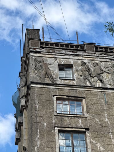 This image shows the facade of a building in Saint Petersburg, Russia, possibly undergoing renovation. The building features bas-relief sculptures of figures, some of which appear to be soldiers holding flags and rifles. The architecture is reminiscent of Soviet-era styles, characterized by its monumental and propagandistic art. The building's exterior shows signs of wear and tear, with crumbling plaster and weathered stone blocks. Several windows are visible, some with reflective glass that captures the sky and clouds. Overhead, electrical wires crisscross against a backdrop of a partly cloudy blue sky, and a branch of a tree is visible in the upper right corner. The building's corner is covered in green netting, suggesting ongoing construction or repair work.