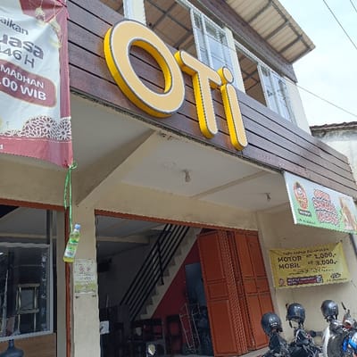 The image shows the exterior of a storefront in Kaliwungu, Indonesia, located at coordinates -6.962 latitude and 110.256 longitude. The storefront has a large sign that reads "OTI" in yellow letters. The storefront is made of wood and has a brown awning. There is a stairway leading to an upper level, and the front entrance is made of double brown wooden doors. There are several motorcycles parked outside. A sign near the motorcycles reads "Tarif Parkir Belanjaan Motor Rp 1.000,- Mobil Rp 3.000,-." A sign on the left wall says "Madhan 10.00 WIB" and a banner that says "Usa 146". A plastic bottle is attached to a green string hanging from the awning above the entrance.