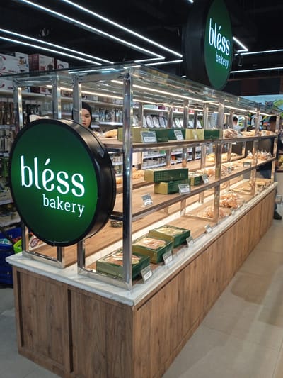 The image depicts a "bléss bakery" display case located inside a building, likely a supermarket or department store, judging by the presence of other retail items in the background. The case is made of wood and metal, with glass shelves containing various baked goods packaged in clear plastic bags. Some goods are also in green boxes. Prices are displayed on small tags. A woman wearing a head covering is partially visible behind the display. The bakery appears to be located in Madiun, Indonesia based on the brand name. The lighting is provided by long, overhead fluorescent lights.