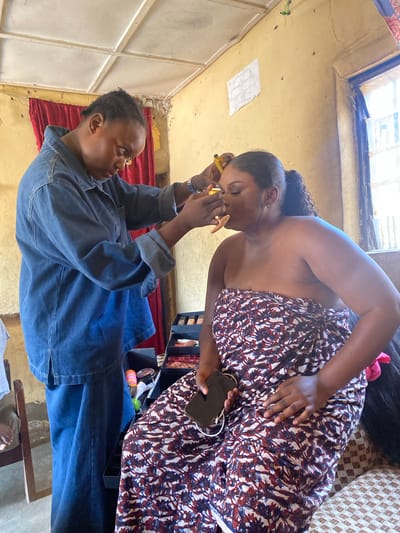 In Panyam, Nigeria, a person in a denim jacket is applying makeup to another person who is seated and wrapped in a patterned fabric. The seated individual has their eyes closed and their face tilted upwards, while the person applying makeup holds a cosmetic tool near their eye. The environment appears to be an indoor space with yellow walls, a white ceiling, and red curtains visible in the background. There is also a window with a view of the outdoors. A makeup kit is open on a table beside the seated person, and a mobile phone rests on their lap.