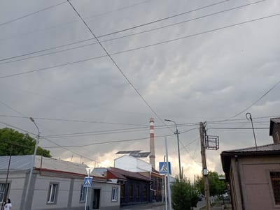 This image depicts a street scene in Gyumri, Armenia, under a heavily overcast sky. The foreground features a paved road and sidewalk. On the left side of the street, there are several buildings with corrugated metal roofs and gray textured walls. One building has a prominent chimney with red and white stripes in the background, suggesting an industrial area or a power plant. Solar panels are visible on the roof of a white building. Various utility poles and a complex network of overhead wires dominate the sky, crossing from multiple angles. Two blue pedestrian crossing signs are visible, one on the left and another on the right side of the street. A woman in a white t-shirt is walking on the sidewalk on the far left. Green trees are present on the left and further down the street on the right. The overall impression is of a somewhat utilitarian urban environment.