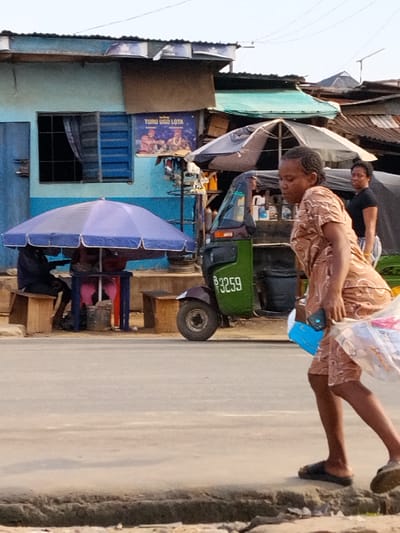 The image captures a street scene in Aba, Nigeria. An adult woman in a brown patterned dress and sandals walks right to left in the foreground. She carries a blue container and a plastic bag. Behind her is a green auto-rickshaw (tuk-tuk) with the number "B 3259" painted on its side. A woman in a black shirt and patterned skirt stands near the tuk-tuk. To the left, a blue building features a window and a sign reading "TURU UGO LOTA." A blue umbrella shelters people seated at a table. The environment includes various corrugated iron roofing structures and street-side stalls. The road occupies a significant portion of the frame.