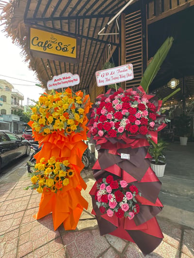 In Nha Trang, Vietnam, a street scene is captured with a prominent yellow sign reading "Cafe Số 1" hanging above a thatched roof structure. In front of the cafe, two large floral arrangements stand on display. The arrangement on the left is primarily composed of yellow roses and sunflowers, wrapped in bright orange paper. A smaller bunch of yellow roses is visible at the base. The arrangement on the right features a mix of red and pink roses, presented in a dramatic arrangement of red and dark brown wrapping. A small white tag is attached to this arrangement. In the background, a black sedan is parked on the left side of the frame. Behind the cafe, trees and more foliage are visible, along with hints of buildings and a glimpse of the sky. The ground is paved with patterned bricks.