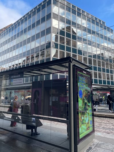 The image depicts an urban scene at the "Alt Winmärik" tram stop in Strasbourg, France. In the foreground, a modern glass and dark-framed tram shelter features an information panel on the left displaying "STRASBOURG Alt Winmärik" and routes for Tram B and F. A digital advertisement on the right pillar shows a vibrant food image with French text. Inside the shelter, a person wearing a dark coat is seated on a bench, facing away from the camera, and another individual in a reddish-orange top stands near the information panel. Beyond the tram tracks, several pedestrians are visible on a sidewalk. The prominent background element is a multi-story building with a reflective glass facade that mirrors a clear blue sky with scattered clouds and the distinct dark-roofed tower of a traditional stone building, along with reflections of other historic architectural structures. Bare tree branches are visible on the far right.