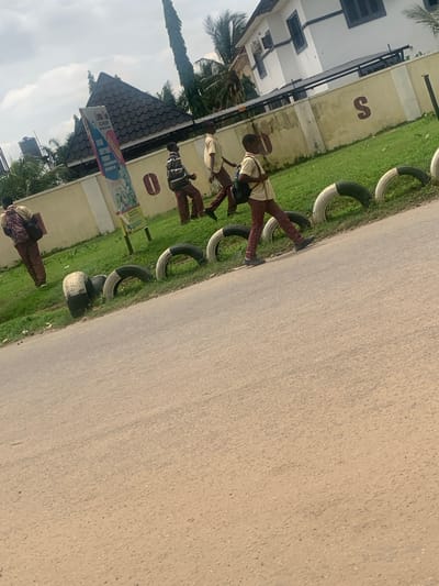 The image depicts an outdoor scene in Abuja, Nigeria, featuring a group of young people walking near a roadside. The environment includes a paved road in the foreground, a grassy area with several repurposed tires used as roadside barriers, and a wall behind that. A building, possibly a school or residence, with white walls and dark window frames, is visible in the background. A tall, slender tree stands to the left of the building. A large banner is posted on a pole near the wall.
There are at least four individuals, appearing to be students due to their attire, which includes light-colored shirts and patterned trousers. They are dressed in what seem to be school uniforms. One individual, on the far left, is seen from the back, carrying a backpack. Three other individuals are walking towards the right side of the frame. One of them is carrying a backpack on their shoulders. The individuals appear to be in motion, walking along the grassy area beside the road. The sky above is overcast with clouds. The overall impression is that of a typical urban or semi-urban street scene in Nigeria.