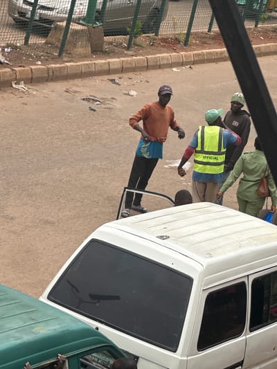 In Bukuru, Nigeria, a group of individuals, appearing to be either workers or bystanders, are gathered on a dusty, outdoor street. In the foreground, the roof of a white van is visible, obscuring the lower portion of the scene. To the left, the roof of a green vehicle is also present. The central focus of the image is on a group of four people in the middle ground. One man, wearing an orange long-sleeved shirt and dark trousers, stands facing the viewer with his hands visible. To his right, another man wears a bright yellow safety vest with the word "OFFICIAL" printed in black letters across the back. He has a green cap on and is wearing light-colored trousers. Behind him, another individual in a camouflage-patterned hat is partially visible. A fourth person, dressed in a green uniform, stands to the right of the man in the yellow vest, holding a brown bag. A small child is partially visible through an open car door. The background consists of a road surface, a concrete curb, and a chain-link fence. Beyond the fence, a car and some vegetation are discernible. The overall environment appears to be a somewhat unkempt urban setting.