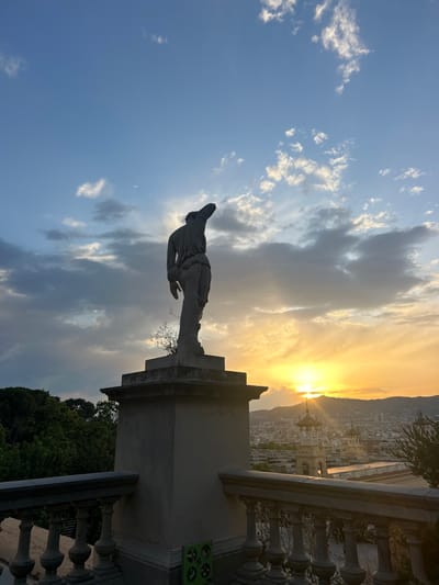 This image captures a sunset over the city of Barcelona, Spain, as seen from the Parc de Montjuïc. In the foreground, a weathered stone statue of a robed figure stands silhouetted against the sky. The statue is mounted on a tiered pedestal, with a small plant growing from its base. A stone balustrade with decorative pillars lines the edge of the viewpoint.

The background is dominated by the warm hues of the setting sun, which casts a golden glow over the city. The sky is a blend of blues, grays, and oranges, with scattered clouds. The city itself stretches out below, a tapestry of buildings with a notable structure featuring ornate domes in the middle distance. Lush green trees are visible on the left side of the image, indicating a park or garden setting. The overall atmosphere is serene and picturesque, highlighting the beauty of Barcelona at dusk.