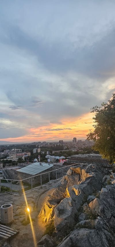 A wide-angle, vertical, eye-level shot captures a panoramic view of Plovdiv, Bulgaria, at sunset from Nebet Tepe, one of the city's ancient hills. The sky is a dramatic canvas of muted blues, grays, and vibrant streaks of orange and pink near the horizon, with wispy clouds scattered throughout. In the foreground, large, rough-hewn rocks dominate the right side of the frame, illuminated by a warm, yellow light emanating from a ground-level fixture on the left. A metal railing runs along the edge of the rocky outcrop.

Below the immediate rocky foreground, there is a paved area with a glass-covered structure and stone ruins. A few people are visible in this middle ground, some standing and observing the view, one possibly playing a guitar. A large red and white sign with "Dr. F" is visible on a building in the distance.

The cityscape stretches out towards the horizon, featuring a mix of modern and older buildings, including several tall structures. Lush green trees dot the urban landscape, softening the architectural lines. The overall impression is one of ancient history meeting a contemporary city under a breathtaking sunset.